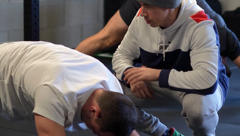 Benjamin, accroupi à côté d'un adhérent en position de gainage, lui corrige sa posture pendant le cours collectif.